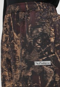 Windbreaker broek in een zwart en bruin abstract patroon. Het materiaal lijkt lichtgewicht met een elastische tailleband. Bevat een "No Problemo" label.