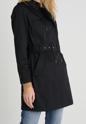 Femme portant un trench-coat noir ceinturé avec des boutons croisés sur un pull beige et un jean bleu.