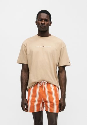 Muž oblečený v béžovom tričku Tommy Hilfiger a oranžových prúžkovaných šortkách so šnúrkou, stojaci pred jednotnou stenou.