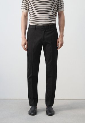 Homme portant un pantalon noir slim, des chaussures noires et une chemise beige à manches courtes avec des rayures horizontales noires, debout sur un sol gris.
