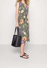 Kwiatowa sukienka na ciemnozielonym tle, z pomarańczowymi i purpurowymi kwiatami, z wiązanym w pasie krojem, zestawiona z czarną torbą na ramię i płaskimi sandałami.