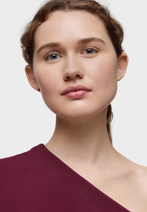 Top burdeos de un solo hombro, tejido texturizado, escote suave. Pendientes en forma de corazón dorados. Maquillaje suave y natural; cabello ligero y despeinado.