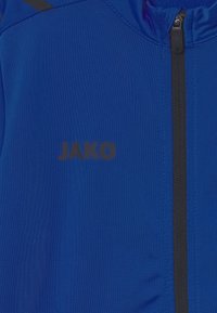 Blaues Sportjackett aus glattem Stoff, mit einem schwarzen Reißverschluss und dem Logo "JAKO" in schwarzer Schrift. Texturiertes, leichtes Design.