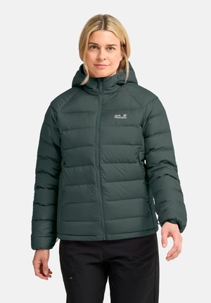 Frau trägt eine dunkelgrüne gesteppte Jacke mit Kapuze und schwarze Hose, steht vor einem einfarbigen hellen Hintergrund.