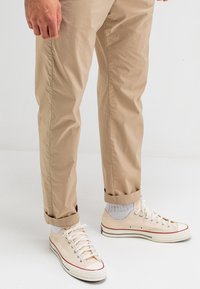 Canvas sneakers in beige met rode en zwarte accenten, voorzien van een vetersluiting en rubberen zool. Gedeeltelijk gedragen met opgerolde lichtkhaki broeken en witte sokken.