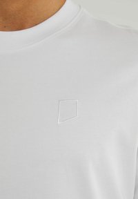 Balta medvilnės marškinėliai su apvaliu iškirpimu, ant krūtinės turi maža siuvinėta geometrinė logotipas, pritaikyta baltai siūlai.