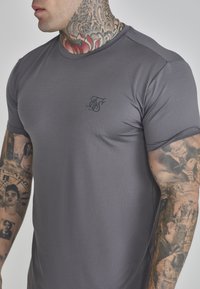 Tricou sportiv gri, realizat dintr-un material texturat. Are mâneci scurte și un logo mic, întunecat pe piept.