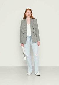 Mulher vestindo um blazer xadrez preto e branco, blusa branca, jeans claros rasgados, tênis brancos, segurando uma pequena bolsa branca, em pé sobre um chão cinza.