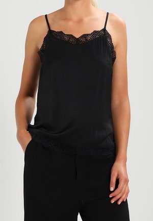 Camisole nera con bordo in pizzo, dotata di spalline regolabili e texture liscia. Abbinata a pantaloni neri in un contesto neutro.