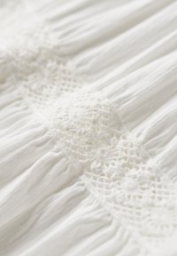 Weißer Stoff mit strukturiertem, plissiertem Design. Die Spitzenborte zeigt aufwendige Blumenmuster und gitterartige Details in der Mitte.