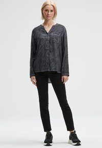 Blonde Frau steht da und trägt ein dunkelgraues, gemustertes Hemd mit Knöpfen, schwarze schmale Hosen und schwarze Sneakers mit weißen Sohlen.