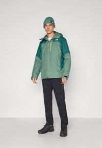 Veste imperméable verte et teal avec capuche, fermeture éclair à l'avant et poches, associée à un pantalon noir et des chaussures de sport noires.