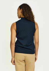 Haut sans manches en tricot bleu marine avec un col haut, texture côtelée au niveau de l'encolure et de l'ourlet, associé à un pantalon beige cintré.