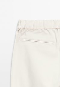 Primo piano di pantaloni beige chiaro con elastico in vita e una singola tasca posteriore orizzontale su uno sfondo bianco semplice.
