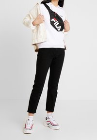 Felpa bianca con tasca frontale, giacca in corduroy color crema, pantaloni neri e sneakers bianche con dettagli rossi e blu navy. Borsa a tracolla nera con logo.