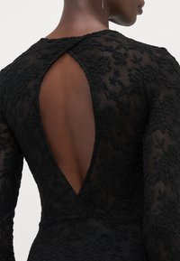 Svarte blondekjole med dyp V-nakke bak, teksturert blomsterdesign, lange ermer og tettsittende silhuett.