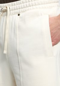 Gros plan sur un pantalon de survêtement crème clair avec une taille élastique, un cordon, une poche avant et une main partiellement dans la poche.