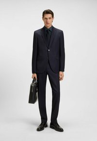 Merenkruunu puku, jossa on räätälöity istuvuus, yhdistettynä pilkulliseen paitaan ja solmioon. Mustat nahkakengät ja tyylikäs musta salkku viimeistelevät asun.