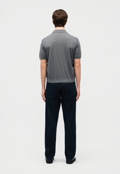 Άνδρας που στέκεται με την πλάτη, φορώντας γκρι πόλο με κοντά μανίκια και σκούρο τζιν μπροστά σε απλό λευκό φόντο.