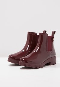 Bottes de pluie bordeaux avec une finition brillante, hauteur cheville, panneaux latéraux élastiques et languettes pour un enfilage facile. Semelle en caoutchouc avec des rainures pour l'adhérence.