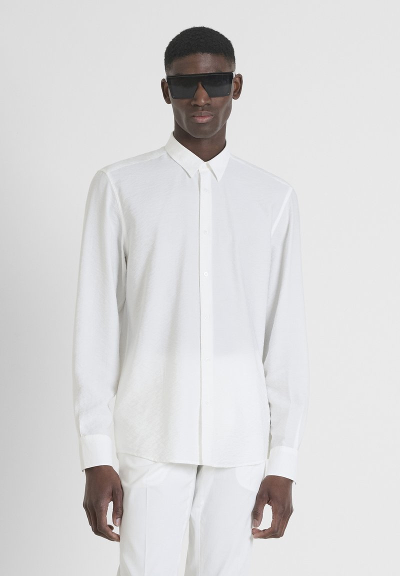 Camicia bianca con colletto a punta, maniche lunghe e un tessuto leggero e testurizzato. Abbinate a occhiali da sole scuri e angolari.