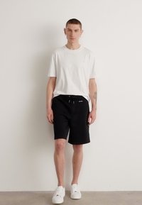 Bílý bavlněný tričko doplněný černými kraťasy na šňůrku. Model má na sobě bílé tenisky. Jednoduchý design, standardní střih, bez vzorů, čisté linie.