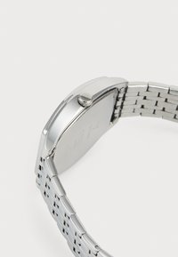 Bracelet et boîtier de montre en métal argenté vus de côté, mettant en avant la couronne et une partie du fermoir à maillons.