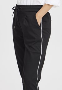 Mustat pehmeästä kankaasta valmistetut jogger-housut, joissa on kiristysnyöri vyötäröllä, sivutaskut ja valkoinen kontrastivetoketju yksityiskohtana lahkeen varrella.
