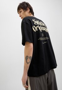 Černé bavlněné tričko s velkým krémovým nápisem "Studio Originals" na zádech, se dalšími potištěnými detaily. Model má na sobě šedé kalhoty.
