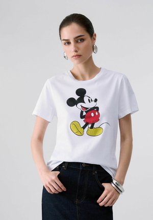 Joven mujer con cabello oscuro lleva una camiseta blanca con estampado colorido de Mickey Mouse y vaqueros oscuros, de pie con las manos en los bolsillos contra un fondo liso.