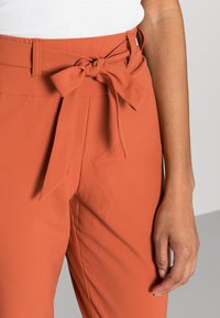 Calças laranja de cintura alta com um laço amarrado na cintura, apresentando uma textura suave e bolsos laterais ocultos.