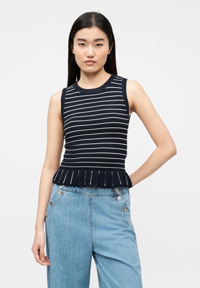 Νέα γυναίκα με μακριά σκουρόχρωμα μαλλιά, φοράει αμάνικη πλεκτή μπλούζα με ναυτικό μπλε και λευκές ρίγες και ανοιχτόχρωμα τζιν ψηλόμεσα παντελόνια.