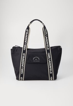 Sac cabas noir avec texte blanc de la marque Karl Lagerfeld sur les poignées, la poche avant et la fermeture éclair en haut, sur un fond uni.