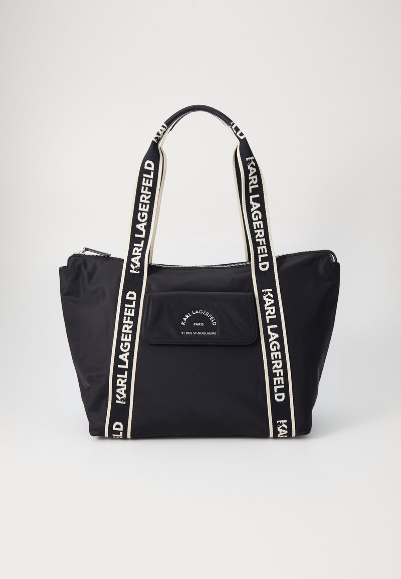 Sac cabas noir avec texte blanc de la marque Karl Lagerfeld sur les poignées, la poche avant et la fermeture éclair en haut, sur un fond uni.