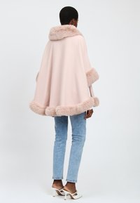 Person iført en lyserød capejakke med pelskrave, lyseblå jeans og hvide hæl-mules, stående med ryggen til på hvid baggrund.