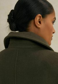 Olive roheline villane mantel kõrge kaelusega ja sooniku detailidega, vaadates tagant. Sile tekstuur, narmas disain ja struktureeritud siluett.