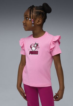 Junges Mädchen mit geflochtenem Haar und Perlen, das ein pinkes T-Shirt mit Rüschensäumen trägt, auf dem die Charaktere von Puma und Hello Kitty abgebildet sind, steht im Profil.