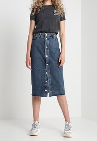 Denimová sukně s knoflíkovým zapínáním vpředu, raw lemováním, tmavě modré barvy a vysokým pasem. V kombinaci se šedým tričkem a bílými teniskami.