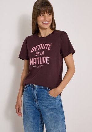Femme souriante aux cheveux bruns porte un t-shirt violet avec le texte "Beauté de la Nature" et un jean bleu, debout devant un fond uni.