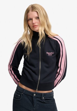 Lyshåret kvinde iført en sort lynlåstrøje med pink striber på ærmerne og "Superdry & Co" logo, kombineret med mørke bukser.