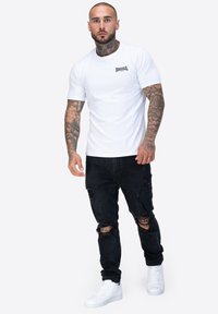 Camiseta blanca de manga corta con un pequeño logo negro, combinada con jeans negros desgastados y zapatillas blancas. La piel está tatuada en los brazos visibles.