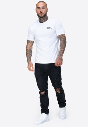 White short-sleeve t-shirt with a small black logo, paired with distressed black jeans and white sneakers. Skin is tattooed on visible arms.