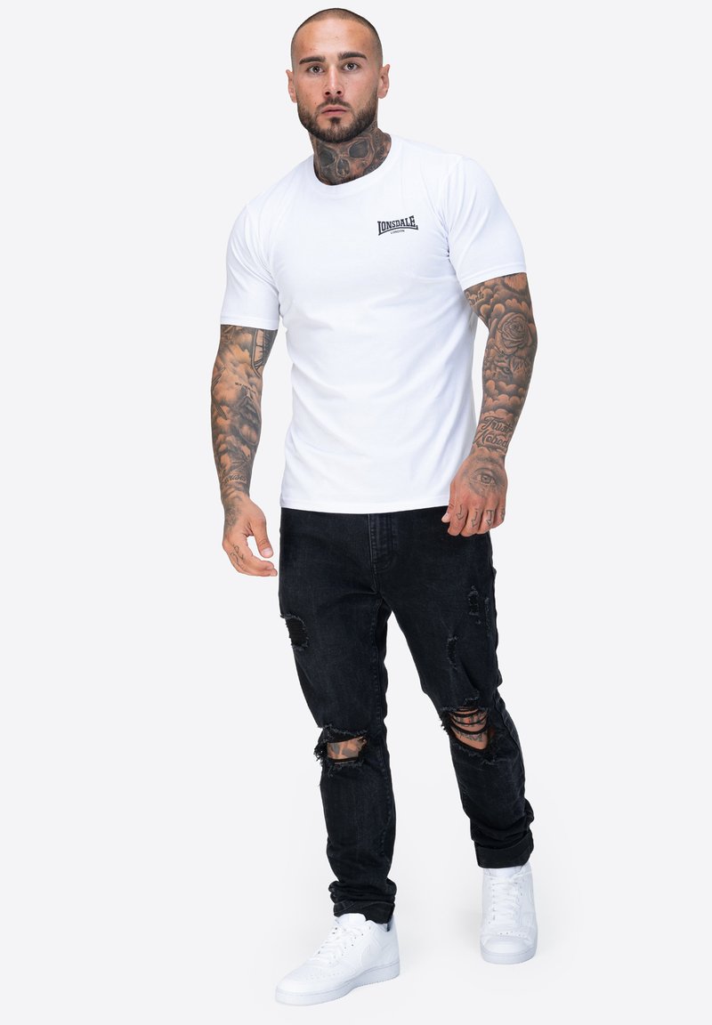 Maglietta bianca a maniche corte con un piccolo logo nero, abbinata a jeans neri strappati e sneakers bianche. La pelle è tatuata sulle braccia visibili.