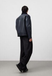 Persona che si allontana indossando una giacca di pelle nera con colletto in shearling, pantaloni neri larghi e scarpe nere su un pavimento grigio.
