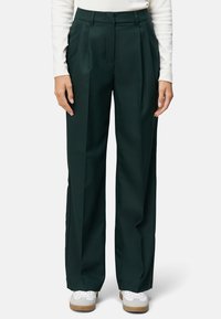 Pantalons plissés taille haute verts foncés à jambe large. Texture lisse, coupe ajustée, sans matériel visible, associés à des baskets blanches.