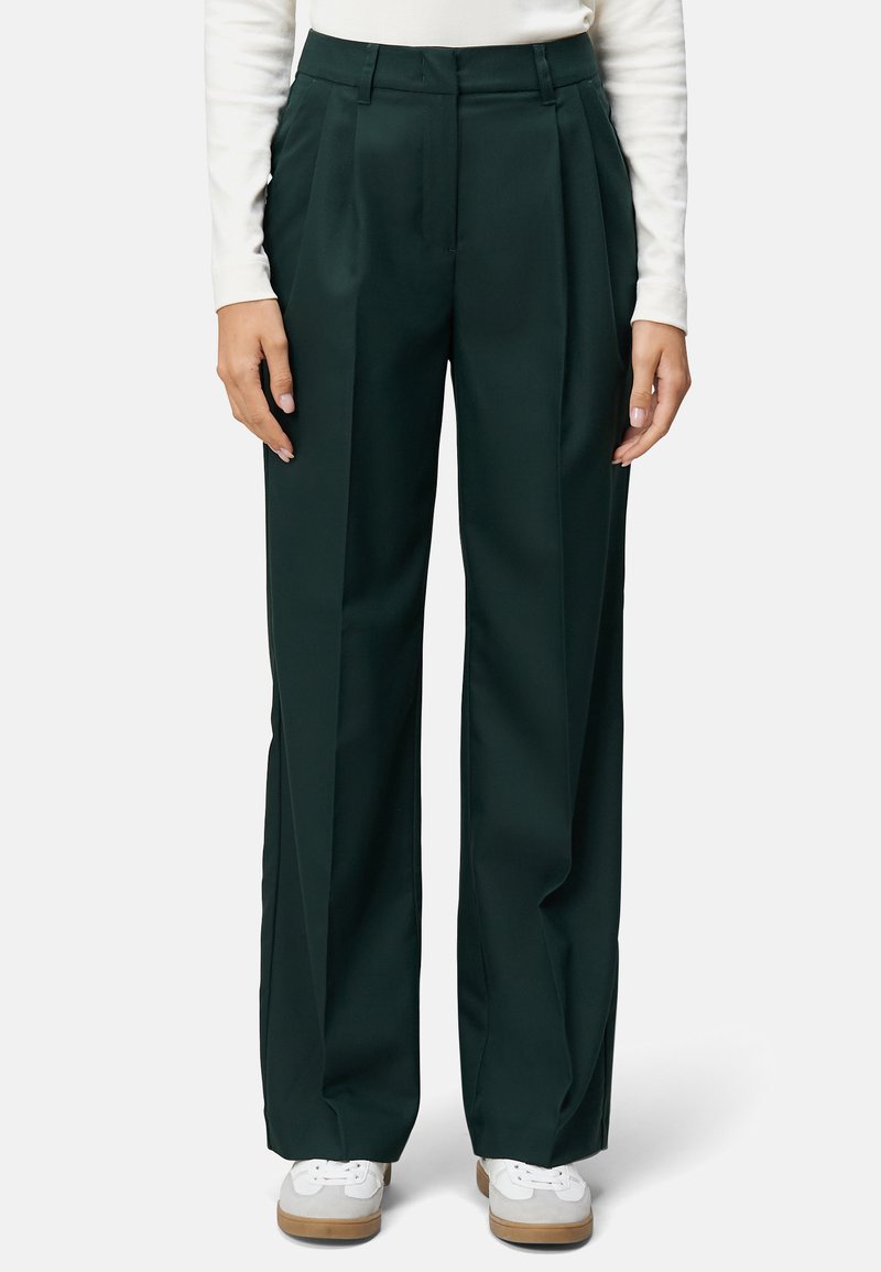 Pantalons plissés taille haute verts foncés à jambe large. Texture lisse, coupe ajustée, sans matériel visible, associés à des baskets blanches.