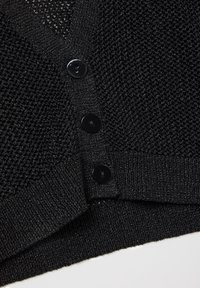 Tissu tricoté noir avec trois boutons noirs sur un vêtement texturé, montrant un gros plan de la zone de fermeture par boutons et de l'ourlet.