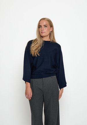 Blonde vrouw met lang haar, gekleed in een marineblauwe trui en grijze wijde broek, staand tegen een witte achtergrond.