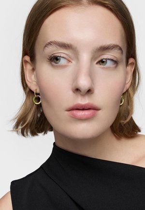 Femme aux cheveux courts châtain clair portant des boucles d'oreilles créoles en or avec des pierres vertes et un haut noir sans manches, regardant sur le côté.