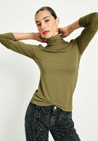 Olivgrön polotröja i mjukt tyg med långa ärmar, stylad med svarta jeans med ormskinnstryck. Slät textur och figurnära design.